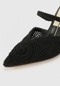 Scarpa nera con punta a forma di freccia, caratterizzata da ricami floreali, una tomaia testurizzata e un design in rete. Include una sottile cinturino alla caviglia per una vestibilità sicura.