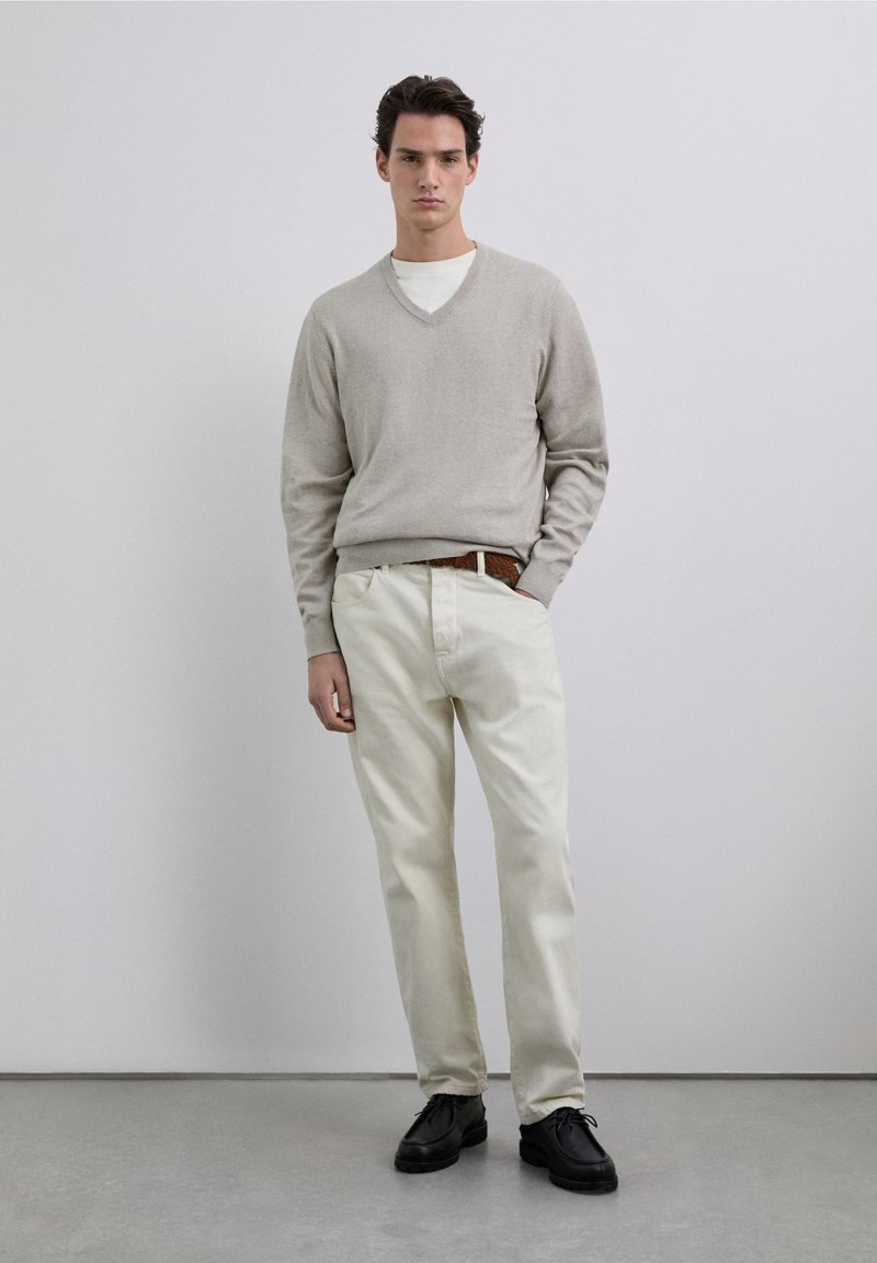 Beiger V-Ausschnitt-Pullover, weißes Hemd darunter, hellcremefarbene Hose und schwarze Schuhe, stehend vor einem neutralen Hintergrund. Detail eines braunen, geflochtenen Gürtels.