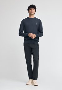Pull en tricot gris foncé avec un col rond, porté avec un pantalon noir slim et des baskets de couleur claire, arborant un petit logo brodé.