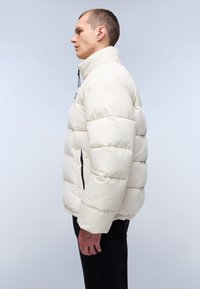 Pufferjack in gebroken wit met een gewatteerd ontwerp, hoge kraag en ritszakken. Kenmerkt elastische manchetten en een iets croppe pasvorm.