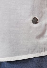 Biały len szew koszuli z małym okrągłym metalowym guzikiem z wygrawerowanym napisem „Marco Polo Denim” nad ciemnoniebieską tkaniną.