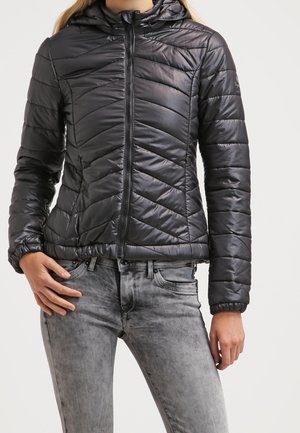 Kvinde iført en sort quiltet hættejakke lynet op, kombineret med falmede grå jeans, stående mod en ensfarvet baggrund.