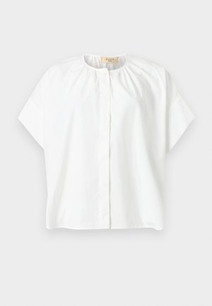 Žena v nosení voľnej bielej blúzky s krátkym rukávom a béžových širokých nohaviciach, drží malú tkanú čierno-bielu kabelku, stojí bosá.