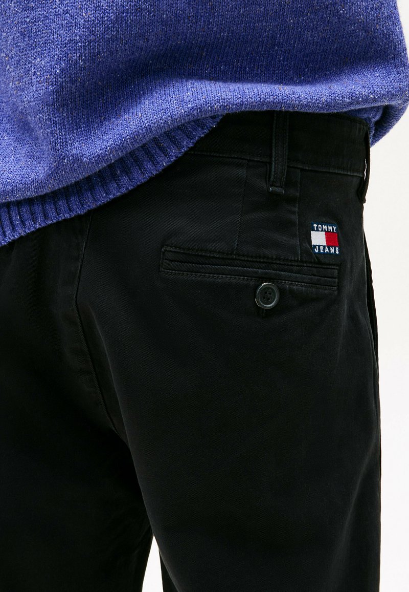 Pantalons noirs avec une texture lisse et une poche arrière arborant un petit logo brodé Tommy Jeans en rouge, blanc et bleu.