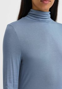 Tops s dolgimi rokavi s turtleneck ovratnikom v svetlo modri barvi, narejen iz mehke tkanine. Ima prilegan obliko in subtilno rebrasto teksturo na ovratniku.