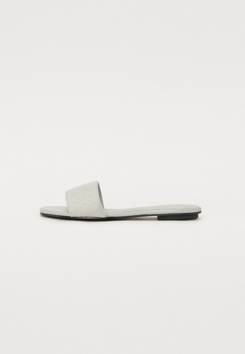 Sandal plat gris avec une large lanière texturée et une semelle lisse. Design minimaliste avec une silhouette basse.