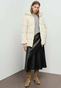 Chaqueta tipo puff en color blanco roto, con diseño acolchado y capucha, combinada con una falda midi de cuero negra y botas de ante marrón claro hasta la rodilla.