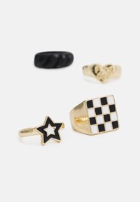 Quatro anéis: um anel preto torcido, um anel em forma de coração dourado, uma estrela dourada com esmalte preto, e um quadrado dourado com padrão quadriculado em preto e branco.