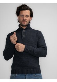 Pulover din tricot de un albastru închis, cu guler înalt și două butoane. Manșete și tiv cu model texturat. Purtați peste blugi albaștri.