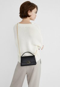Čierna kožená crossbody taška s horným úchytom, zlatým kováním a textúrovaným povrchom; nosená cez krémový pletený sveter a svetlošedé nohavice.
