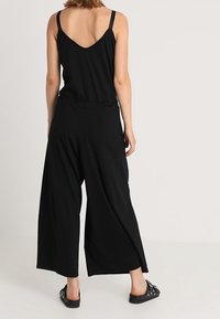 Zwarte wijde jumpsuit met dunne bandjes, voorzien van een v-hals achterkant en een ontspannen pasvorm. Gemaakt van zachte stof, heeft het een rimpel taille detail.
