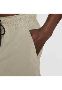 Pantalones cortos beige con cinturilla elástica y cordón, que cuentan con bolsillos laterales y una textura de tela suave.