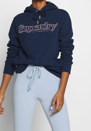 Woman wearing a navy blue Superdry hoodie with logo text and light blue drawstring sweatpants against a white background.