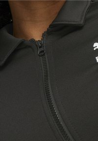 Chaqueta negra con cremallera, fabricada en un tejido texturizado, con un cuello alto y un detalle de logo en blanco en el pecho, cerca de la línea de la cremallera.