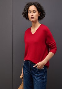 Maglione rosso a scollo a V realizzato in materiale morbido, abbinato a jeans blu scuro. Il maglione ha una vestibilità comoda e polsini a costine. Il modello ha una mano in tasca.