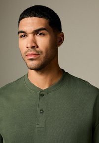 Green, short-sleeve henley shirt made of smooth cotton with a two-button placket and subtle texture, featuring a rounded collar.