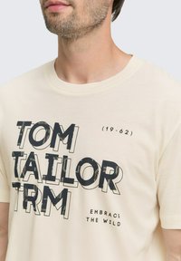 Vīrietis, kurš valkā krēmkrāsas T-kreklu ar melnu tekstu, kas uzrakstīts "TOM TAILOR TRM", "19-62" un "EMBRACE THE WILD" priekšpusē, izgriezts ap kaklu un piedurknēm.