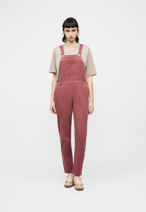 Ung kvinde stående iført falmet lyserøde fløjlsdessiner, beige oversize t-shirt og beige sneakers mod en ensfarvet hvid baggrund.