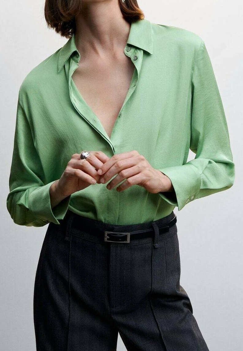 Personne portant une chemise vert clair boutonnée avec un pantalon noir et une ceinture, mains jointes et ornées d'une grosse bague en argent.