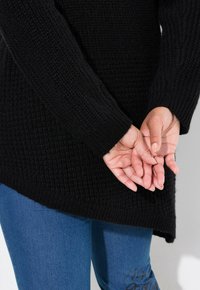Maglione nero a maglia texturizzata con maniche lunghe e orlo asimmetrico, abbinato a jeans in denim blu con un motivo decorativo su una gamba.