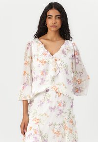 Blouse florale en tissu léger et transparent, avec un col en V à volants, des manches en cloche, et un fond blanc orné de fleurs orange et violettes.