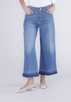 Femme portant un jean bleu à taille haute et jambes larges, avec braguette à boutons et ourlets effilochés, assorti à des ballerines blanches à bride arrière ajourée.