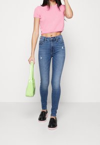 Mujer vestida con un top corto rosa, jeans ajustados azules, mocasines negros con suelas rosas, sosteniendo un pequeño bolso verde a su lado.