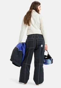 Pantalones de nieve negros con costuras blancas, dos bolsillos traseros, combinados con un pulóver crema y una chaqueta azul, casco en la mano.