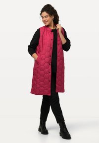 Gepolsterte, lange Weste in Fuchsia mit hexagonalem Steppdesign, mit Druckknöpfen, zwei Seitentaschen und ohne Kragen, kombiniert mit schwarzen Hosen.