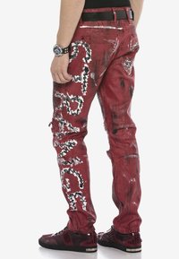 Rote, abgenutzte Jeans mit weißen und schwarzen grafischen Mustern, schmaler Schnitt und seitlichen Taschen. Getragen mit schwarzen, Nieten besetzten Schuhen.
