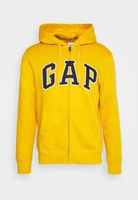 Sudadera amarilla con cremallera y capucha ajustable con cordón, logo de GAP en azul marino, bolsillos delanteros y puños acanalados. Fabricada con una tela suave de textura lisa.