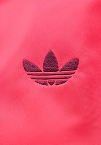 Logo Adidas trèfle brodé violet foncé sur un tissu texturé rose vif.