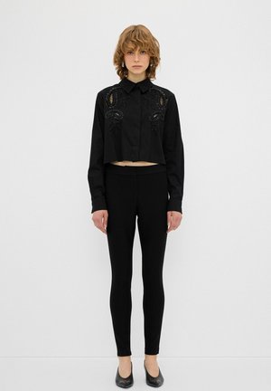 Sort cropped skjorte med intrikationssort broderi, parret med sorte leggings og flade sorte sko. Simpelt design, mat stofstruktur.