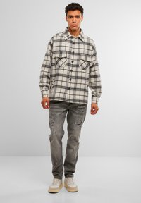 Cămașă din flanel în carouri, crem și negru; are două buzunare pe piept, închidere cu nasturi și mâneci lungi. Poartă cu blugi gri cu aspect uzat și adidași.