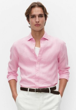 Nuori henkilö, jolla on vaaleanruskeat hiukset, käyttää vaaleanpunaista napitettavaa paitaa valkoisen aluspaidan päällä, valkoiset housut ja kudottu tummanruskea vyö, kädet taskuissa.