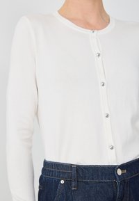 Biały kardigan z długim rękawem i okrągłym dekoltem, zapinany na pięć błyszczących guzików, w połączeniu z ciemnoniebieskimi dżinsami.