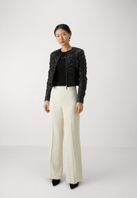 Chaqueta negra texturizada con cierre de cremallera, combinada con una blusa negra ajustada y pantalones anchos color crema, llevados con zapatos de tacón negros.