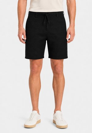 Mann trägt schwarze Shorts mit Kordelzug, ein helles Shirt und weiße Sneaker, stehend vor schlichtem, hellem Hintergrund.