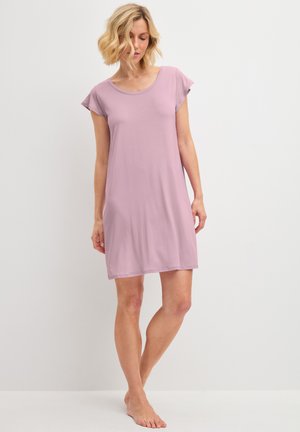 Frau mit kurzen blonden Haaren, die ein locker sitzendes, hellviolettes, knielanges Kleid mit kurzen Ärmeln trägt, barfuß stehend.