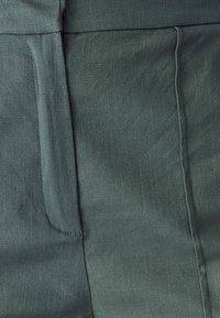 Zelená ľanová látka s jemnou textúrou, vidite�ľným prešívaním a detailom vrecka na boku, s jemným, svetlým leskom.