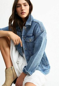 Giacca di jeans azzurro chiaro con chiusura a bottoni, due tasche sul petto e vestibilità comoda, abbinata a un vestito bianco e stivaletti beige alla caviglia.