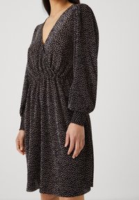 Schwarzes Kleid mit tiefem V-Ausschnitt, langen Puffärmeln und taillierter Passform. Besitzt ein strukturiertes Muster aus kleinen, hellen Punkten.