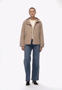 Beige huvad jacka med knappar framtill, sidofickor och en något överdimensionerad passform, i kombination med blå vida jeans och svarta stövlar.