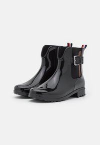 Bottines noires brillantes avec une semelle en caoutchouc, des panneaux latéraux élastiques et une boucle décorative. Présente des accents rayés rouge, blanc et bleu.