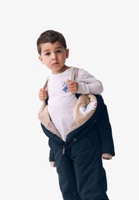Blaues Kapuzenjacke mit beigem Innenfutter, offen. Graues Langarmshirt mit einem kleinen Grafikdruck. Das Kind passt die Jackenriemen an.