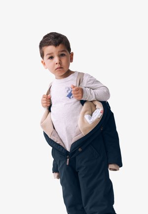Blaues Kapuzenjacke mit beigem Innenfutter, offen. Graues Langarmshirt mit einem kleinen Grafikdruck. Das Kind passt die Jackenriemen an.