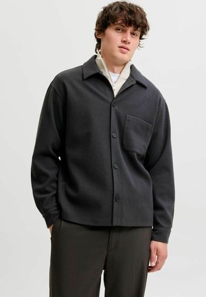 Jeune homme aux cheveux bouclés portant une veste sombre boutonnée sur un haut clair zippé, posant avec une main dans la poche devant un fond uni.