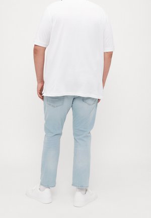 Personne portant une chemise blanche à manches courtes, un jean bleu clair, des chaussettes blanches et des baskets blanches, debout devant un fond uni.
