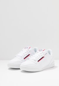 Weiße Sneaker mit einem glatten Obermaterial aus Leder, strukturierten Einsätzen, rotem Akzentstreifen und einem Logo an der Seite. Schnürsenkel und gepolsterter Kragen für zusätzlichen Halt.