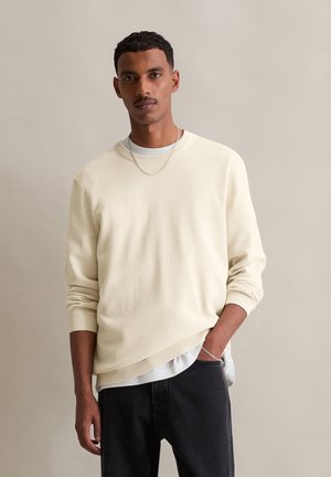 Pull en tricot beige clair avec une texture côtelée subtile, coupe décontractée et col rond, superposé à une chemise blanche, associé à un jean foncé.